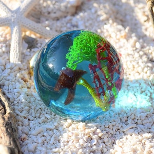 May include: A clear glass sphere filled with blue water, green and red coral, and a brown seahorse. The sphere is sitting on a bed of white sand.