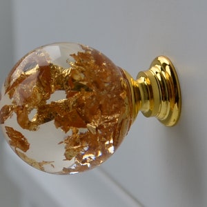 May include: A clear glass cabinet knob with gold leaf flakes embedded inside. The knob has a gold metal base.