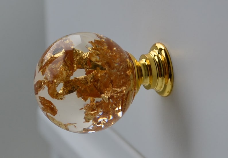 May include: A clear glass cabinet knob with gold leaf flakes embedded inside. The knob has a gold metal base.