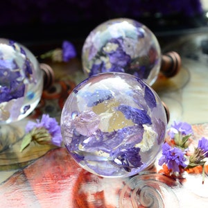 May include: Clear glass cabinet knobs with dried purple flowers encased inside. The knobs have a round shape and are attached to a bronze-colored base. The background features more purple flowers.