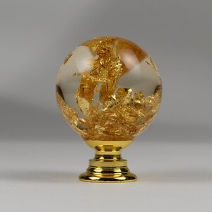 May include: A gold and clear glass cabinet knob with a round, globe-shaped design. The knob is filled with gold leaf flakes and has a gold metal base.