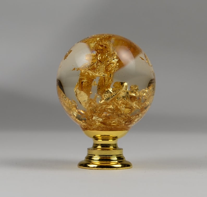 May include: A gold and clear glass cabinet knob with a round, globe-shaped design. The knob is filled with gold leaf flakes and has a gold metal base.