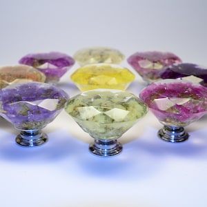 以下が含まれることがあります： 銀色のベースが付いた、ダイヤモンド型のキャビネットノブ9個セット。各ノブには、紫、ピンク、黄色のさまざまな色合いの押し花が詰められています。