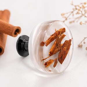 Puede incluir: Un pomo de armario ovalado transparente con palitos de canela en su interior, sujeto a una base negra. Palitos de canela y anís estrellado se disponen cerca sobre una superficie blanca, con flores secas en el fondo. Este pomo decorativo añade un toque natural.