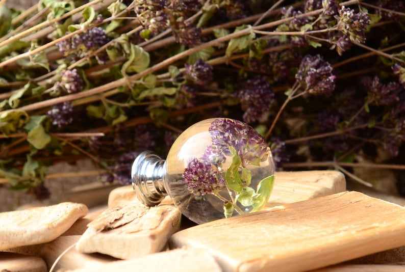 May include: A clear glass knob with a silver base, featuring dried purple and green flowers encased in resin. The knob is resting on a piece of wood.