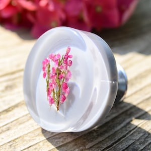 May include: A white round knob with a small sprig of pink flowers pressed into the centre. The knob has a silver metal base.