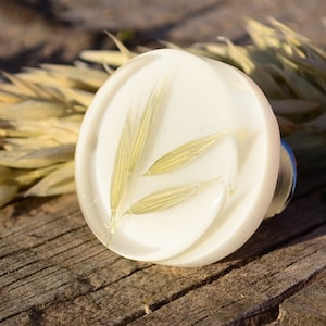 Op de afbeelding: Een witte ronde harsring met een geperst gedroogd havertakje erin. De ring ligt op een houten oppervlak.