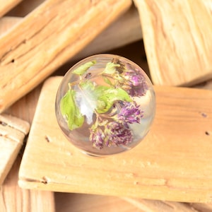 May include: A clear glass sphere containing dried purple flowers and green leaves. The sphere is resting on a piece of wood.