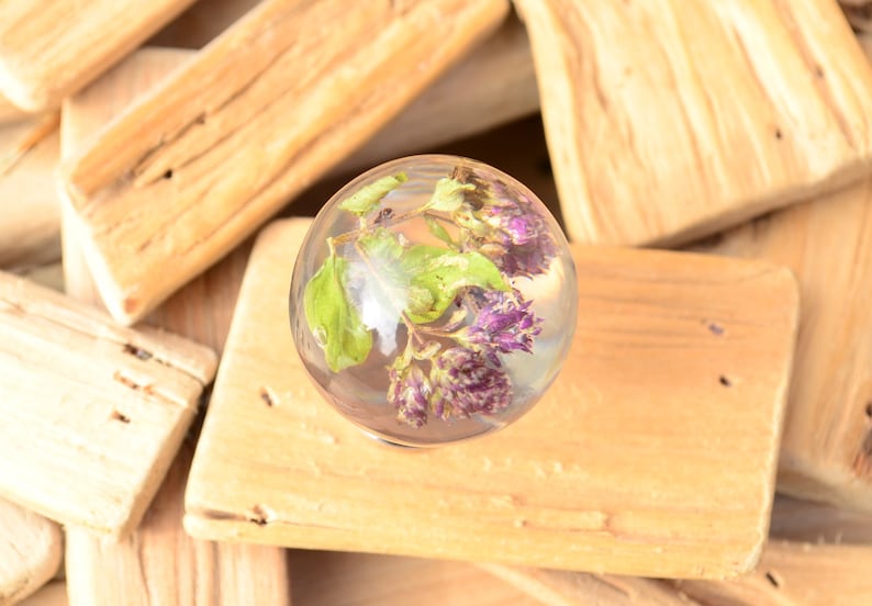 May include: A clear glass sphere containing dried purple flowers and green leaves. The sphere is resting on a piece of wood.