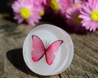 May include: A white round resin cabochon with a pink butterfly design. The butterfly has a dark brown body and pink wings.