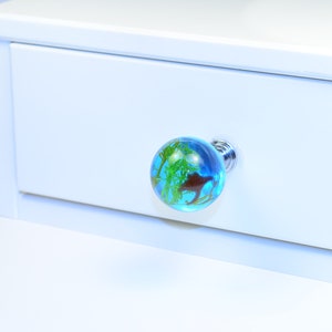 May include: A blue glass drawer knob with a green and brown design. The knob is attached to a white drawer.