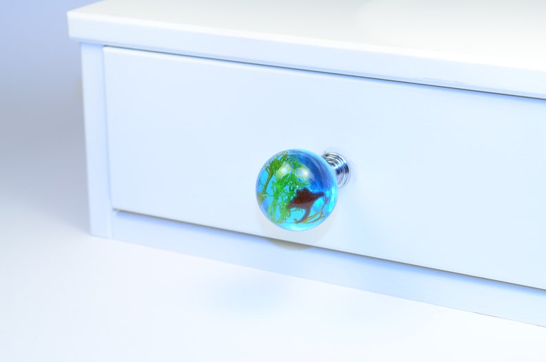 May include: A blue glass drawer knob with a green and brown design. The knob is attached to a white drawer.