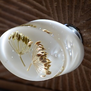 Może przedstawiać: Biały, okrągły gałka drzwiowa z suszonymi kwiatami osadzonymi w przezroczystej żywicy. Kwiaty są jasnobrązowe i mają delikatny, pierzasty wygląd.