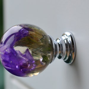 Peut inclure: Un bouton de porte rond en verre transparent avec une fleur violette pressée à l'intérieur. Le bouton est fixé à une base en métal argenté.