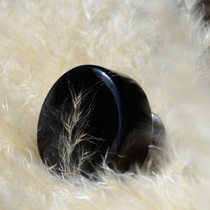 Può includere: Un tappo per bottiglia rotondo nero con un fiore secco pressato all'interno. Il tappo è su una superficie bianca e soffice.