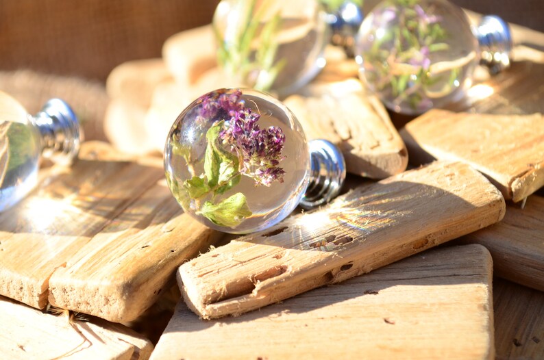 May include: Close-up of a set of clear glass cabinet knobs with dried flowers and greenery encased in resin. The knobs have a silver metal base and are displayed on a rustic wooden surface.