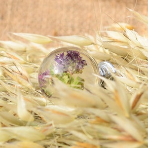 May include: A clear glass sphere containing a small purple flower and green leaves, resting on a bed of dried beige grass.