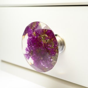 Könnte beinhalten: Ein dekorativer Knopf aus klarem Harz mit einer violetten und goldenen Blume. Dieser Knopf ist perfekt, um Möbeln einen Hauch von Natur zu verleihen.