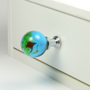 May include: A blue and green globe-shaped drawer pull with a chrome base. The pull is attached to a white drawer.