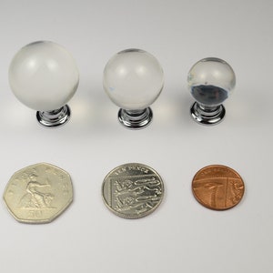 May include: Three clear glass cabinet knobs with chrome bases. The knobs are different sizes and are shown next to three British coins for scale.