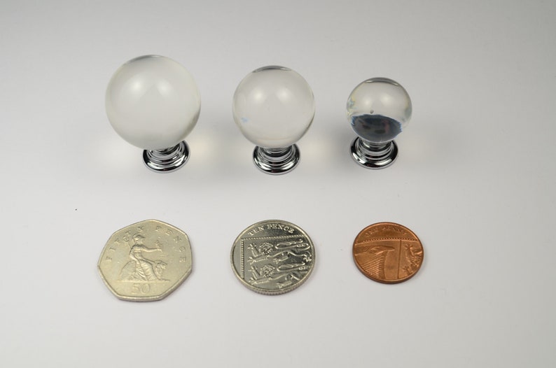 May include: Three clear glass cabinet knobs with chrome bases. The knobs are different sizes and are shown next to three British coins for scale.