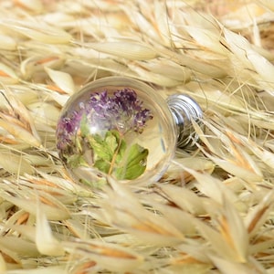 May include: A clear glass sphere with a silver metal base containing a preserved purple flower and green leaves. The sphere is resting on a bed of dried, light brown grass.