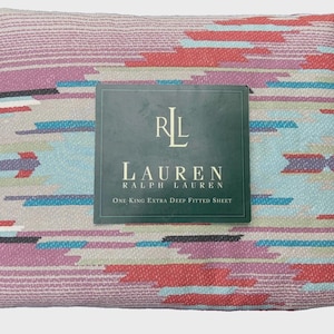 Pode incluir: Um lençol de baixo extra profundo dobrado, tamanho king, com um padrão geométrico em tons de rosa, vermelho, azul e cinza. O lençol tem uma textura tecida e apresenta uma etiqueta com o logotipo "RL LAUREN".