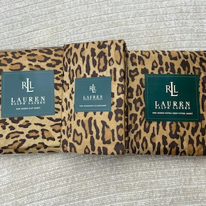 Könnte beinhalten: Drei gefaltete Bettwäsche-Sets mit Leopardenmuster. Die Sets umfassen ein Bettlaken, zwei Standard-Kissenbezüge und ein tiefes Spannbetttuch. Jedes Set hat ein dunkelgrünes Etikett mit dem Markennamen "LAUREN RALPH LAUREN". Die Bettwäsche ist braun und beige.