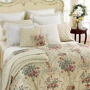 Puede incluir: Una cama con cabecero color crema y ropa de cama floral. La ropa de cama presenta un fondo crema con un estampado de flores rosas y verdes en jarrones. Varias almohadas blancas con bordes con volantes y una manta color crema completan el look.