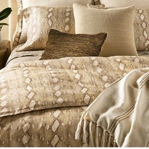 Puede incluir: Una cama con un edredón, fundas de almohada y cojines con estampado de piel de serpiente en tonos beige y marrón. Una manta crema con flecos y rayas marrones oscuras está sobre la cama. La ropa de cama crea una estética neutra y natural.