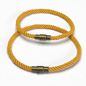 May include: Two gold rope bracelets with a brass clasp. The bracelets are stacked on top of each other.