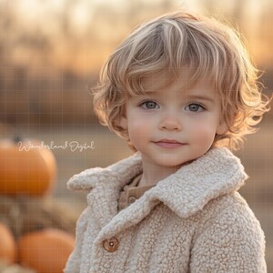 May include: A young child with blonde curly hair wearing a cream coloured fleece jacket. The child is looking at the camera and smiling. The background is blurred and out of focus, with a pile of pumpkins in the background.