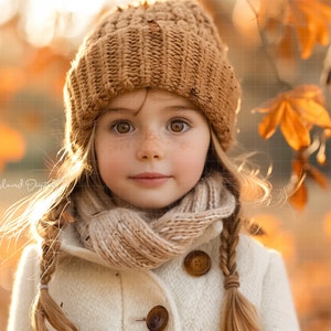 May include: A young girl with long blonde hair wears a brown knitted hat and a cream-colored scarf. She is wearing a white coat with a button closure. The background is a blurred image of autumn leaves.