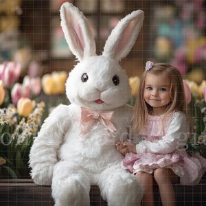 May include: A young girl in a pink dress sits with a large white plush bunny. The bunny has a pink bow around its neck. The girl is smiling and holding the bunny's paw. They are sitting in front of a backdrop of colorful flowers.