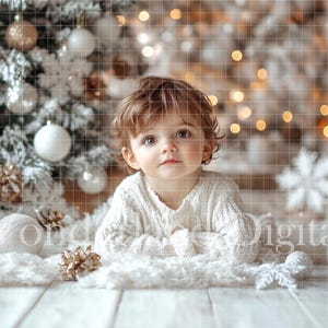 May include: A young child wearing a white sweater sits in front of a white Christmas tree with white and gold ornaments. The child is looking at the camera.