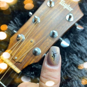 May include: Close-up of a fingernail with a dancer silhouette design. The nail has a glittery, iridescent finish. A wooden guitar headstock with the brand name "Taylor" is in the background.
