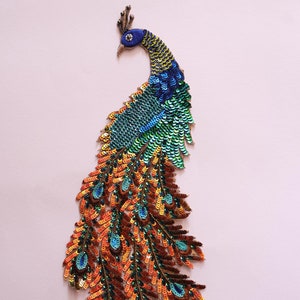 Könnte beinhalten: Eine farbenfrohe Pfauen-Applikation mit schillernden Pailletten. Der Pfau hat einen blauen Kopf und Hals, einen grünen Körper und orangefarbene und grüne Schwanzfedern.