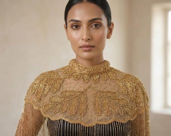 Puede incluir: Una mujer con una capa dorada con cuentas, cuello alto y diseños de hojas. La capa está sobre una blusa negra con rayas doradas verticales. La capa tiene flecos y está hecha de un material transparente.