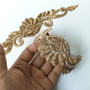 Op de afbeelding: Twee goudkleurige, sierlijke geborduurde applicaties. Eén heeft een gebogen, vleugelachtige vorm en de andere een lang, vloeiend ontwerp. Beide hebben ingewikkelde details met een bloemen- en bladerpatroon en kleine witte kralen.