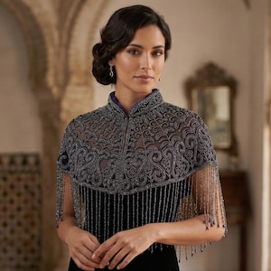 Può includere: Un capelet di perline argentate con ricami intricati e dettagli di frange. Il capelet presenta un colletto alto e una sovrapposizione trasparente, creando un look elegante e sofisticato. Indossato sopra un abito nero.