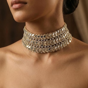 Puede incluir: Un collar gargantilla dorado con un diseño en capas. El collar presenta un patrón repetitivo en forma de hoja con pequeños adornos transparentes. La joya se lleva alrededor del cuello, mostrando sus intrincados detalles y artesanía.