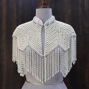 May include: A white beaded cape with a high collar and a chevron design. The cape features long beaded fringe along the bottom edge. The garment is displayed on a mannequin against a dark wood background.