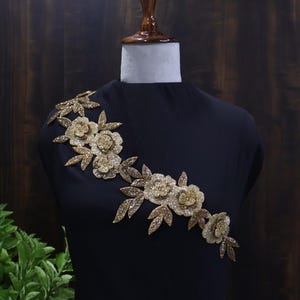 Puede incluir: Vestido negro exhibido en un maniquí, adornado con un elaborado adorno floral dorado sobre el hombro. El intrincado diseño presenta flores y hojas detalladas, creando una estética lujosa. El fondo es de madera oscura.