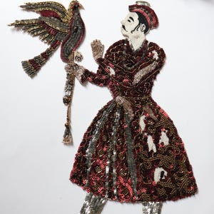 May include: An embroidered applique depicting a figure in a maroon and silver outfit, holding a bird. The applique is decorated with sequins and beads. The bird has maroon, gold, and silver details.