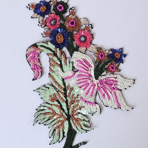 Op de afbeelding: Een bloemvormige applicatiepatch met gedetailleerd kraal- en paillettenwerk in tinten roze, blauw, groen en wit. Het ontwerp bevat gestileerde bloemen en bladeren met een opvallend wit en roze vlinderachtig element in het midden.