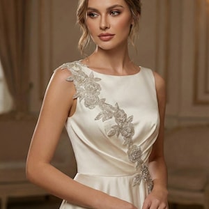 Puede incluir: Vestido sin mangas de color marfil con adornos florales. El vestido presenta un cuello redondo y un corpiño ajustado que fluye hacia una falda amplia. Detalles florales plateados con cuentas caen por el hombro y cruzan el corpiño.