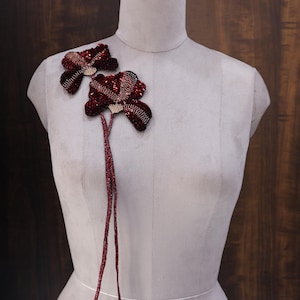 Puede incluir: Dos broches florales burdeos con tallos de cuentas están prendidos a un maniquí. Los broches tienen un diseño en capas con una mezcla de texturas y colores, incluyendo rojo oscuro y acentos brillantes. El maniquí es de color gris neutro.