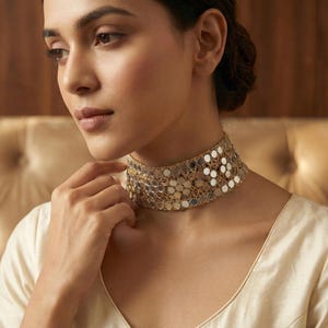Puede incluir: Un collar gargantilla dorado con un diseño de espejo. El collar está hecho de pequeñas piezas redondas y reflectantes, creando un efecto brillante. El collar se lleva en el cuello de una persona, y el fondo es de un color suave y neutro.
