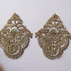 May include: Two ornate, gold-colored embroidered appliques. Each applique features a symmetrical design with floral and leaf motifs, embellished with sequins. The appliques are ideal for embellishing garments or craft projects.