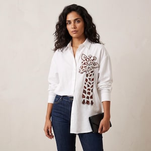 Puede incluir: Una camisa blanca de manga larga con botones y una gran aplicación de cuentas de jirafa en color marrón y blanco en el lado izquierdo. La camisa tiene un cuello clásico, una tapeta de botones frontal, vaqueros azul oscuro y un bolso de mano negro.